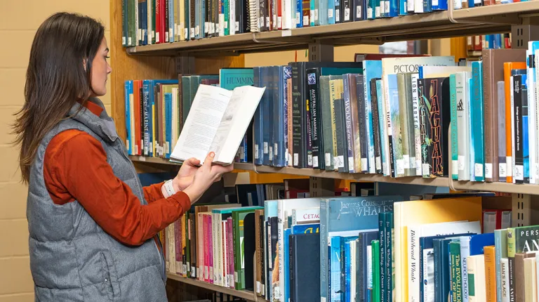 A student stands with an open book in front of shelves of numerous books in a library stack