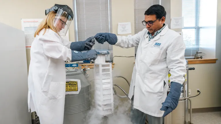 A professor assists a student wearing a protective face mask to pull a container out of cryogenic storage