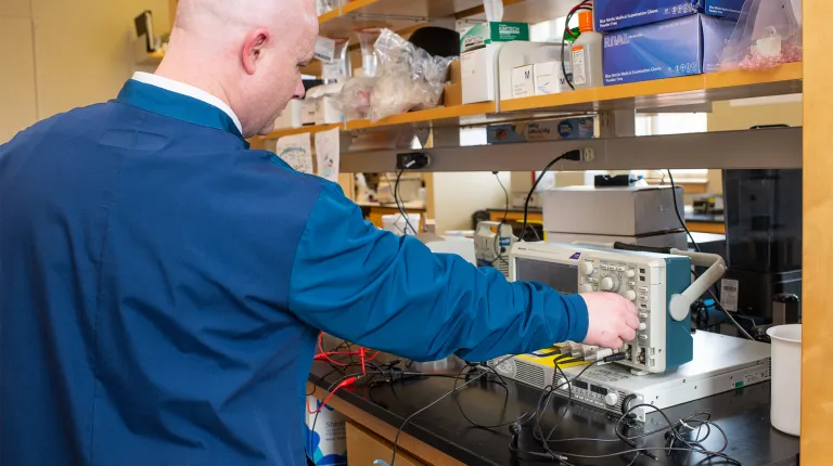A professor turns a knob on an oscilloscope in a laboratory setting