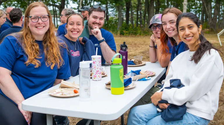 A gorup of six U N E employees sitting at a table during a barbecue smile for the camera