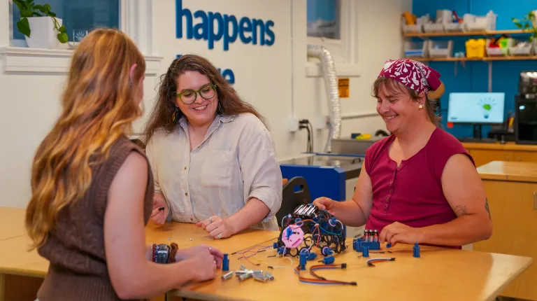 Jessica Howard, Sophia Crockett-Current, and Milo Lypps collaborate on projects at a table beneath "innovation happens here" signage in a makerspace.