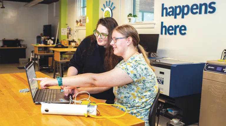 Sophia Crockett-Current and Lillian Westerberg collaborate at a laptop in a makerspace with "innovation happens here" displayed on the wall behind them.