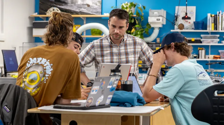 An instructor in a plaid shirt works with three students gathered around a table in a makerspace with bright blue walls.