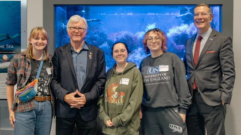 Shaw Innovation Fellows stand with David Shaw and U N E President James Herbert in front of a "Biomedical Exploration in the Sea" display featuring an aquarium backdrop