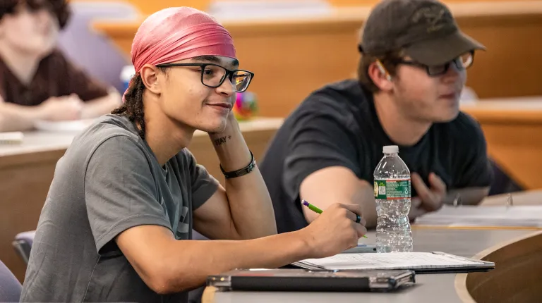 Students engaged in classroom discussion, with one student in a pink headwrap and glasses smiling while taking notes at their desk.