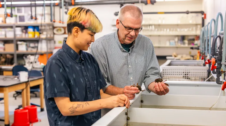 Professor and student examine marine specimens in a laboratory workspace filled with research equipment and storage shelves.