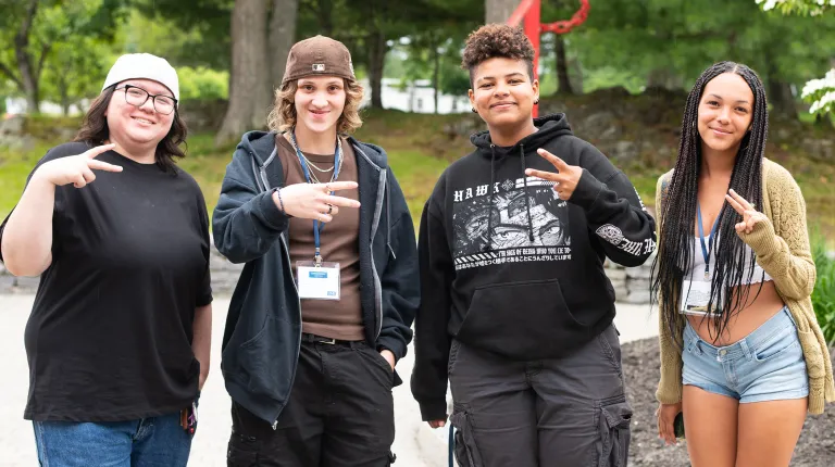 Four students pose together outdoors on campus, making peace signs and smiling for the camera.