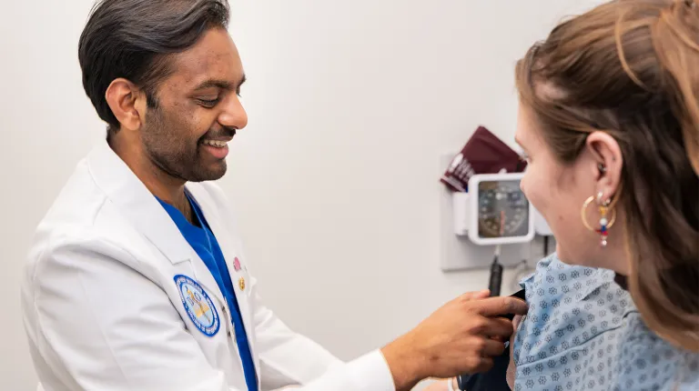 A smiling UNE College of Osteopathic Medicine student in a white coat uses a stethoscope to examine a standardized patient in a clinical practice center exam room.