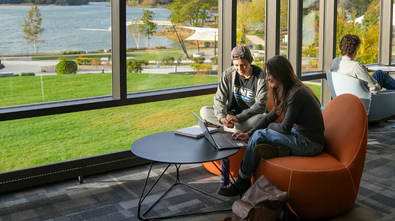 Two UNE students collaborate on a laptop in a modern campus lounge, with floor-to-ceiling windows overlooking the waterfront and autumn foliage.