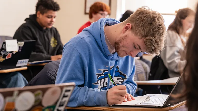 A UNE student in a blue UNE hoodie takes notes in a classroom, surrounded by fellow students working at their desks.