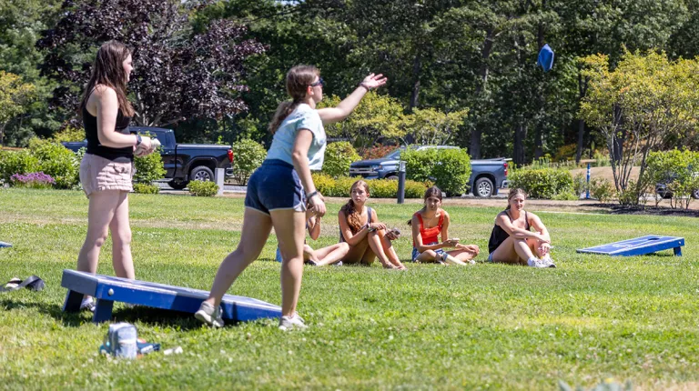 Students play cornhole on a sunny campus lawn during an orientation event, while others sit in the grass watching nearby.