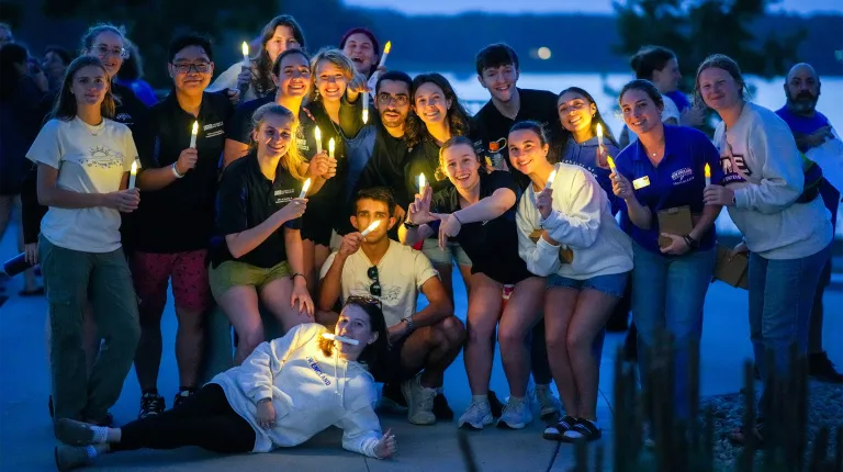 A large group of smiling students hold glowing candles together at dusk near the water during a First Night campus event, several wearing UNE gear.