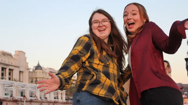 Two students pose enthusiastically with outstretched arms on a European street at dusk with historic buildings in the background.