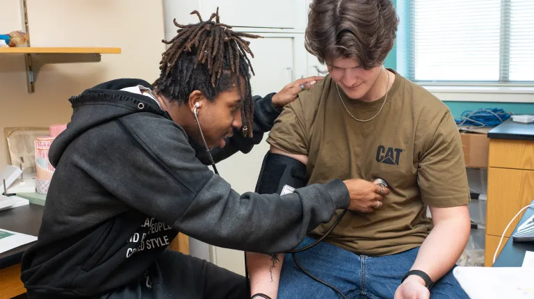 A biology student in a black hoodie takes the blood pressure measurement on a student seated at a desk in a class lab.