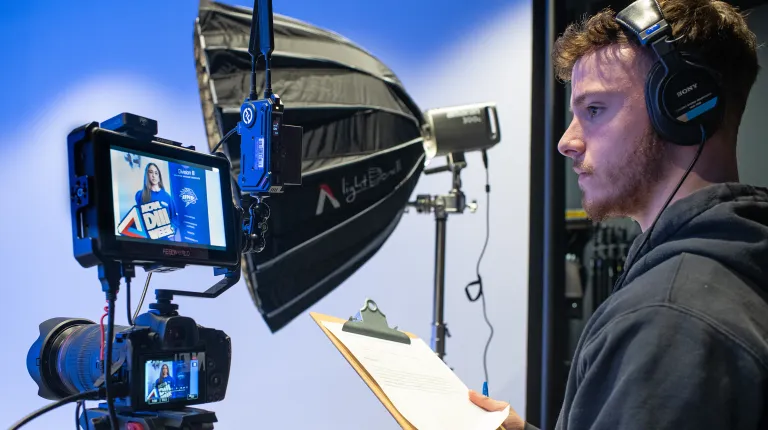 A Communication and Media Arts student wearing headphones reviews a script on a clipboard while monitoring camera equipment with professional lighting and a softbox in a studio setting.