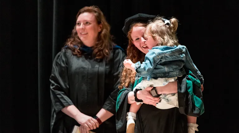 A graduate in cap and gown holds her young daughter on stage during a U N E commencement ceremony while another graduate looks on smiling.