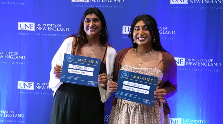 Two U N E College of Osteopathic Medicine students smile in front of a U N E backdrop, holding signs announcing they matched in psychiatry at UMass Chan.