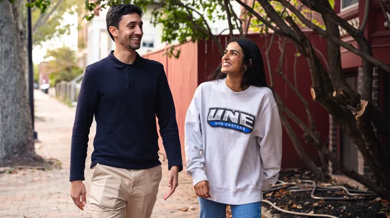 Two students walk and chat along a brick sidewalk in downtown Portland, one wearing a U N E Nor'easters sweatshirt.