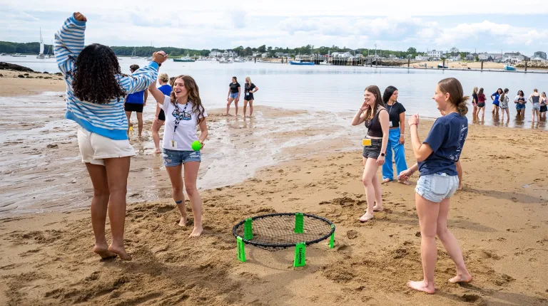 U N E students laugh and play spikeball on Ferry Beach, with the harbor and boats in the background.