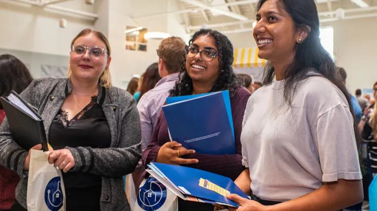 Three U N E College of Dental Medicine students smile while holding informational folders at a career fair.