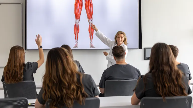 A physician assistant professor points to an image of leg muscles on a large monitor as several seated students raise their hands