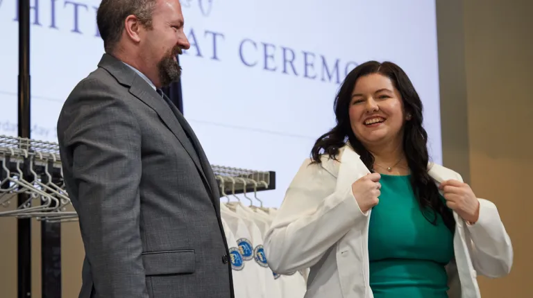A U N E Physician Assistant student smiles while being helped into her white coat at the White Coat Ceremony.