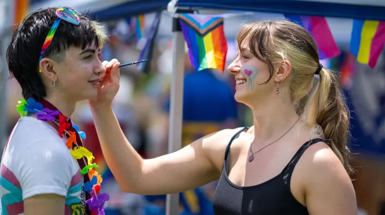 A U N E student paints a heart design on an attendee's face at Portland Pride, surrounded by rainbow flags.