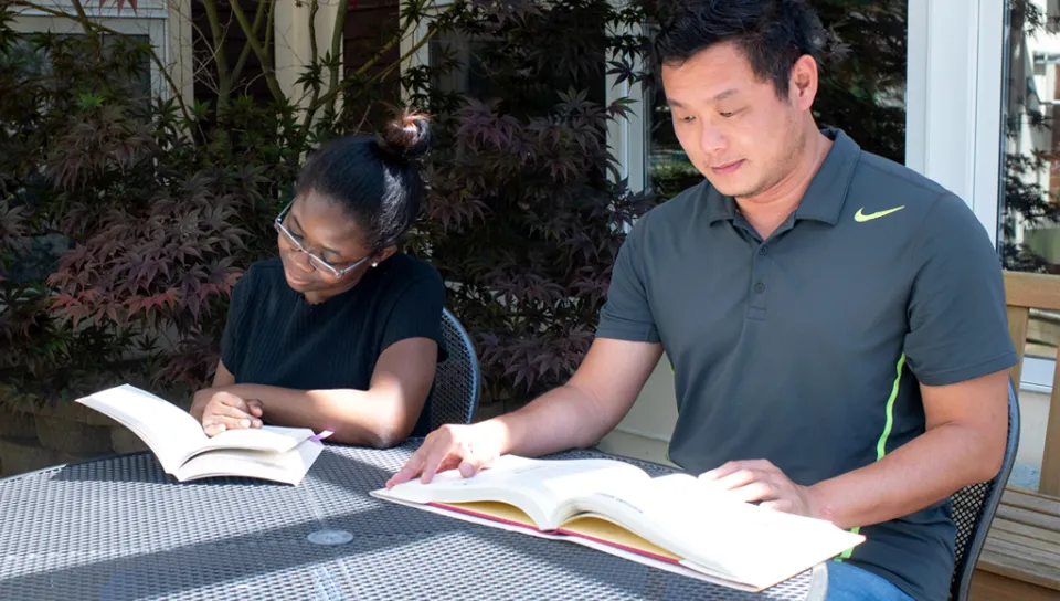 Two U N E students sit together at an outside table each reading a textbook