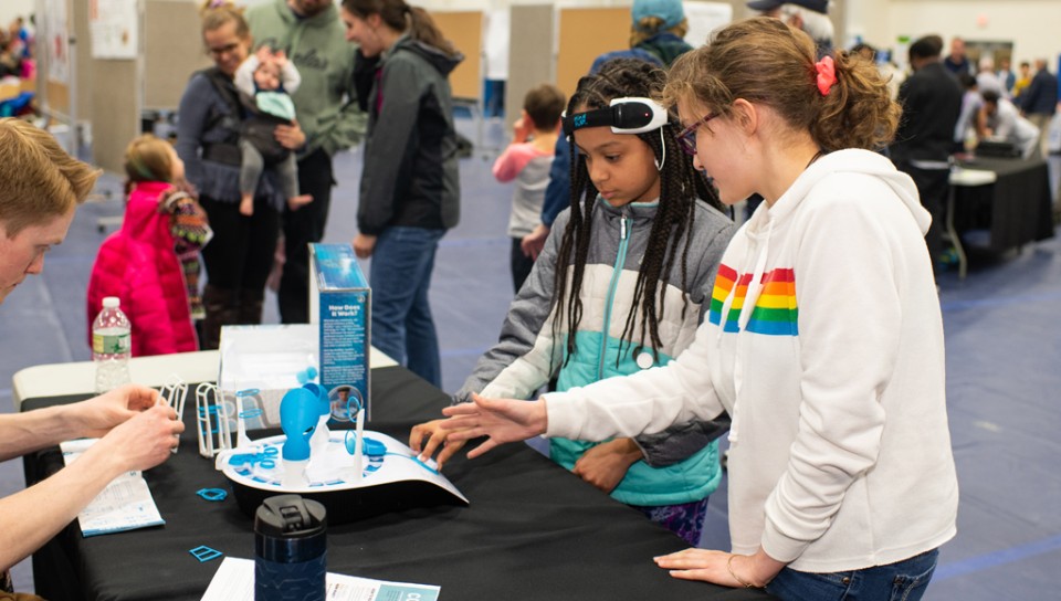 A child wearing scientific equipment learns about its use with her friend at a U N E Brain Fair