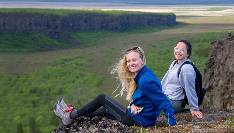 Two students sitting on the edge of a cliff overlooking cliffs and the ocean in Iceland