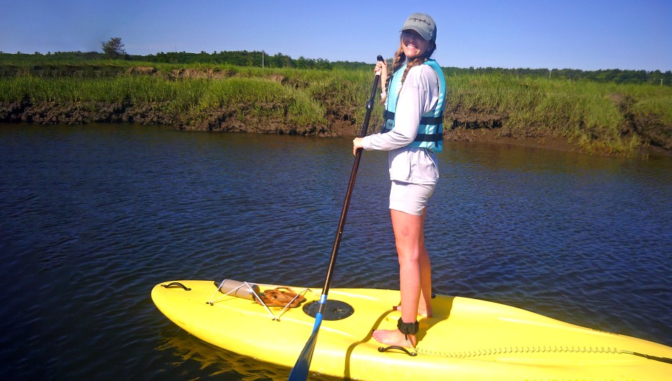 A student smiles at the camera while paddle boarding