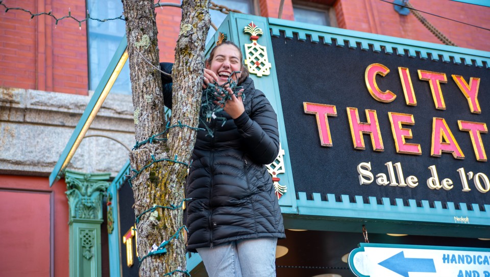 A female student wraps lights around the trunk of a tree in front of Biddeford's City Theater