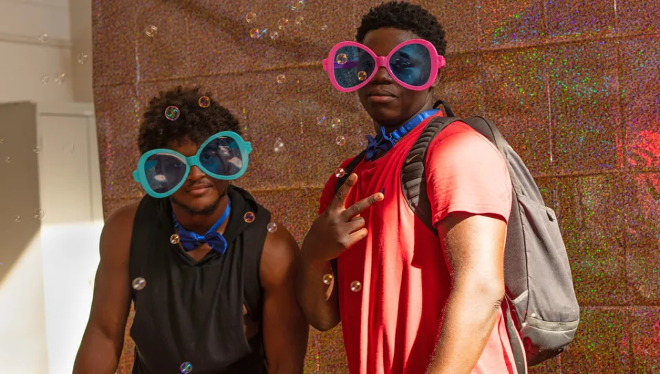 Two students take a photo together while wearing oversized sunglasses in front of a glitter background