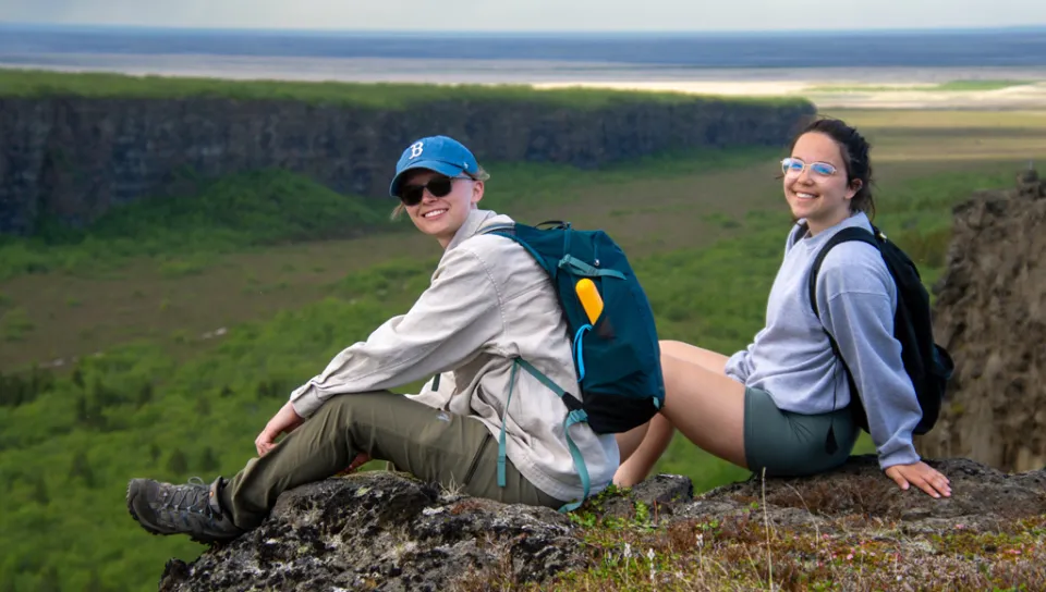 Two U N E students sit on top of a cliff overlooking a green canyon in Iceland