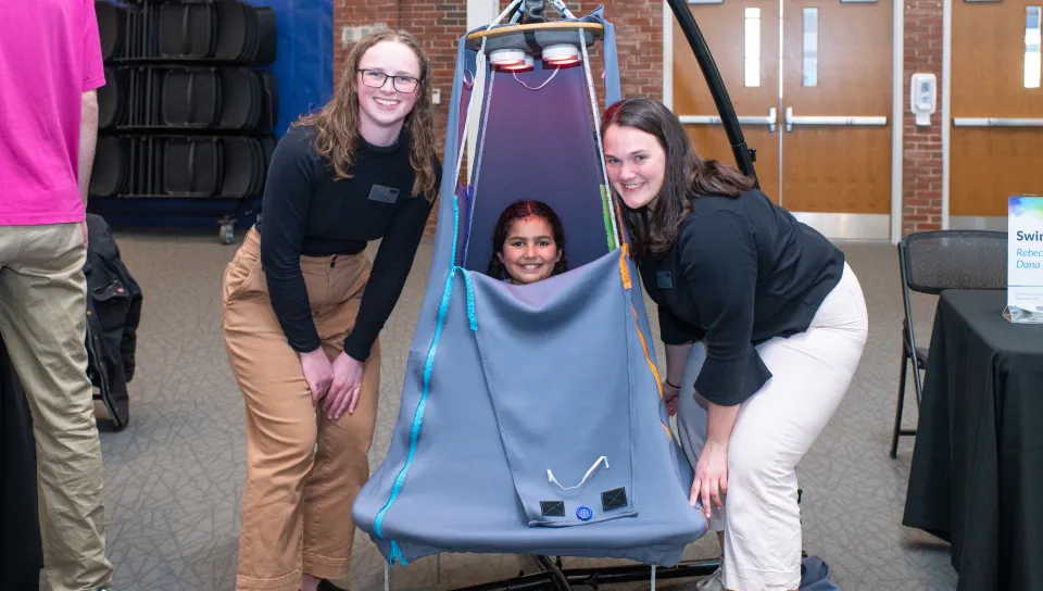 A child sits inside a suspended tent and two students pose around her