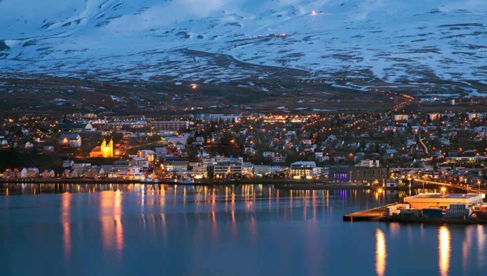 A cityscape at night that sits on the edge of water with snow-covered mountains in the background