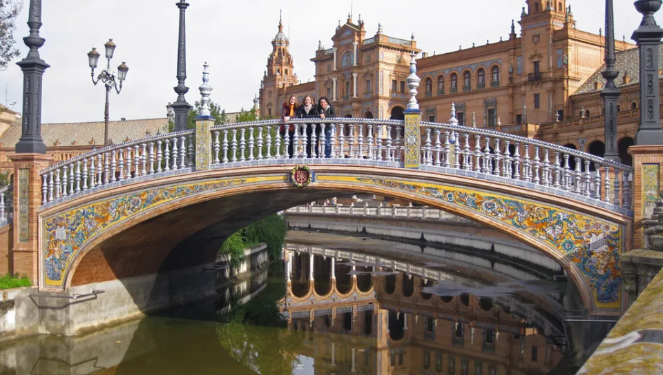 Three U N E students stand on the middle of a bridge over a river surrounded by Spanish architecture
