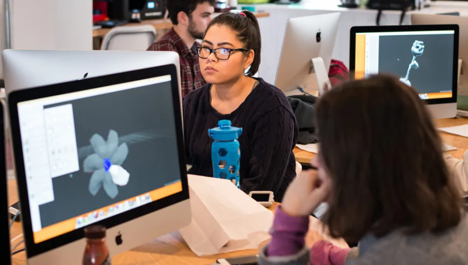 Two computer monitors display 3D renderings being worked on by U N E students