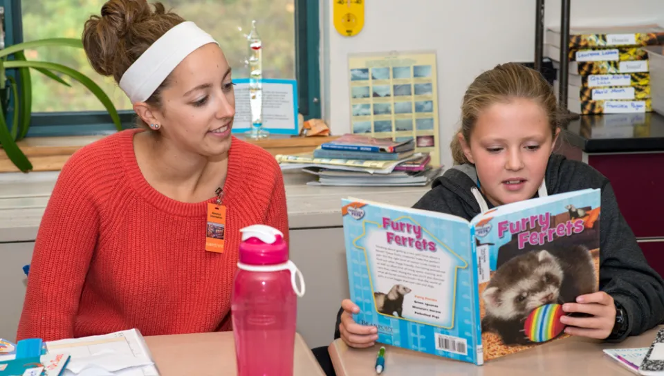 An education student sits with a middle school student as she reads from a book titled, "Furry Ferrets"