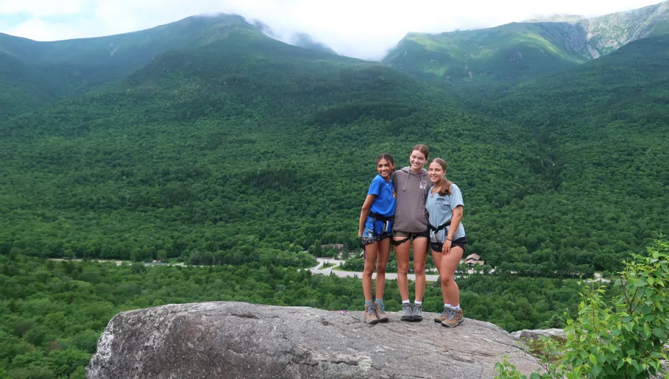 Three students stand on a rock in front of green mountains