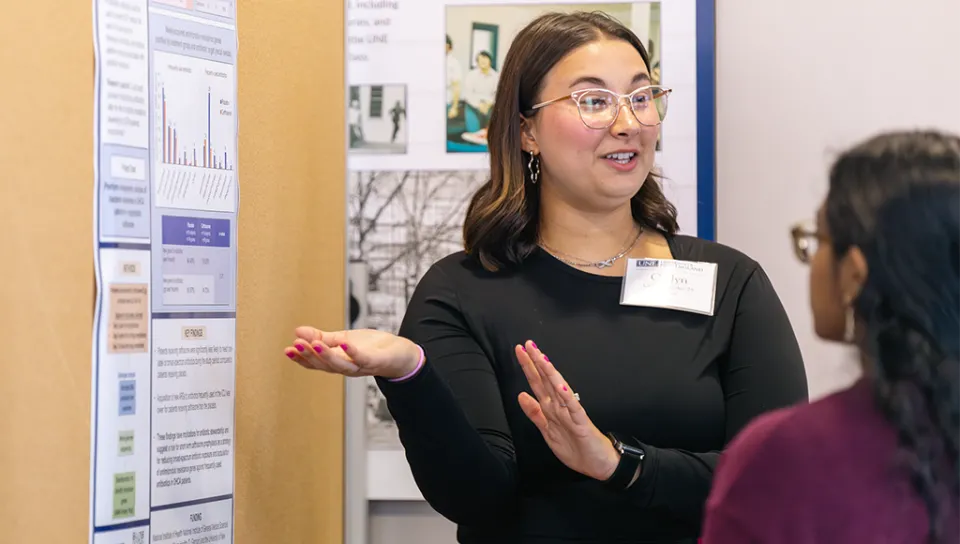 A U N E student gestures at a poster while explaining her research to a few attendees of a fall research symposium.