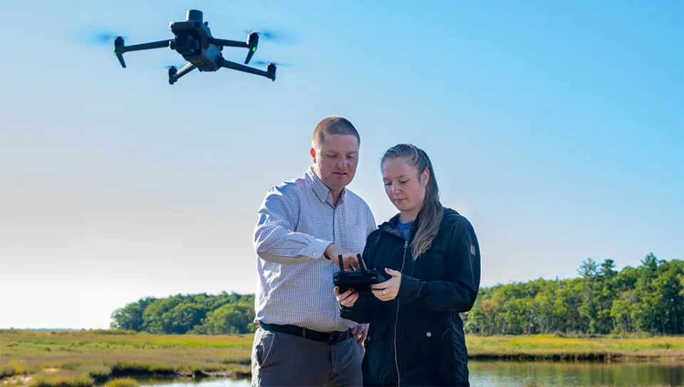 Will Kotchtitzky points to drone while Katie DeWater flys drone over megapools in a marsh next to the Biddeford Campus.