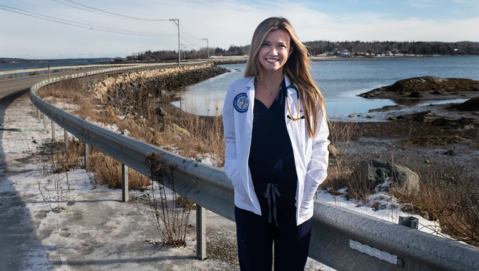 A C O M student in their white coat stands in front of the Maine coast