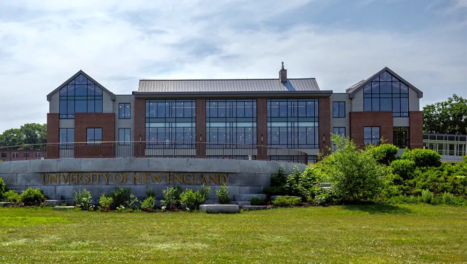 Brick building with large glass windows sits behind a stone sign that reads "University of New England" on a summer day