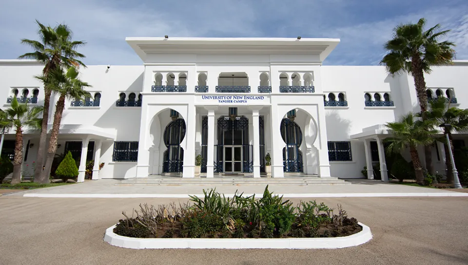 A white building with columns surrounded by a flower bed and palm trees, under a clear sky featuring a few clouds.