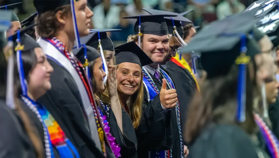 Two people in academic regalia smile and give the camera a 'thumbs up' signal from the student crowd at a commencement ceremony