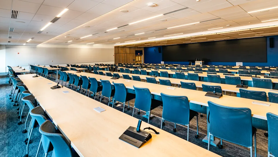 A large monitor extending most of a wall sits behind several rows of light-wood tables and blue chairs