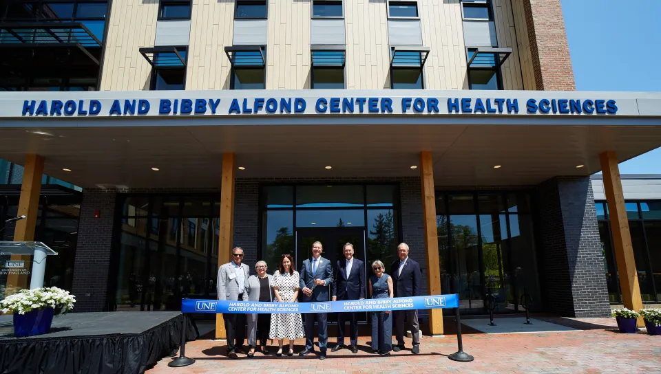 UNE administrators and trustees pose in front of a UNE-branded ribbon in front of the Harold and Bibby Alfond Center for Health Sciences