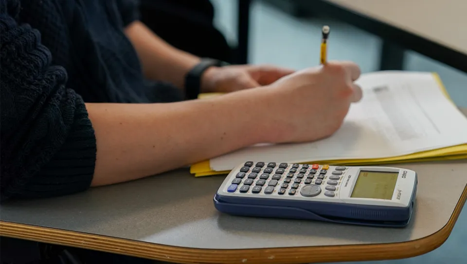 A student uses a pencil to write on a sheet of paper with a calculator next to them on their desk