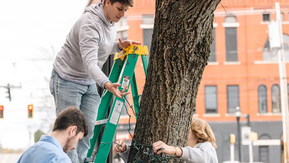 Three U N E students wrap string lights around a small tree in front of a brick building
